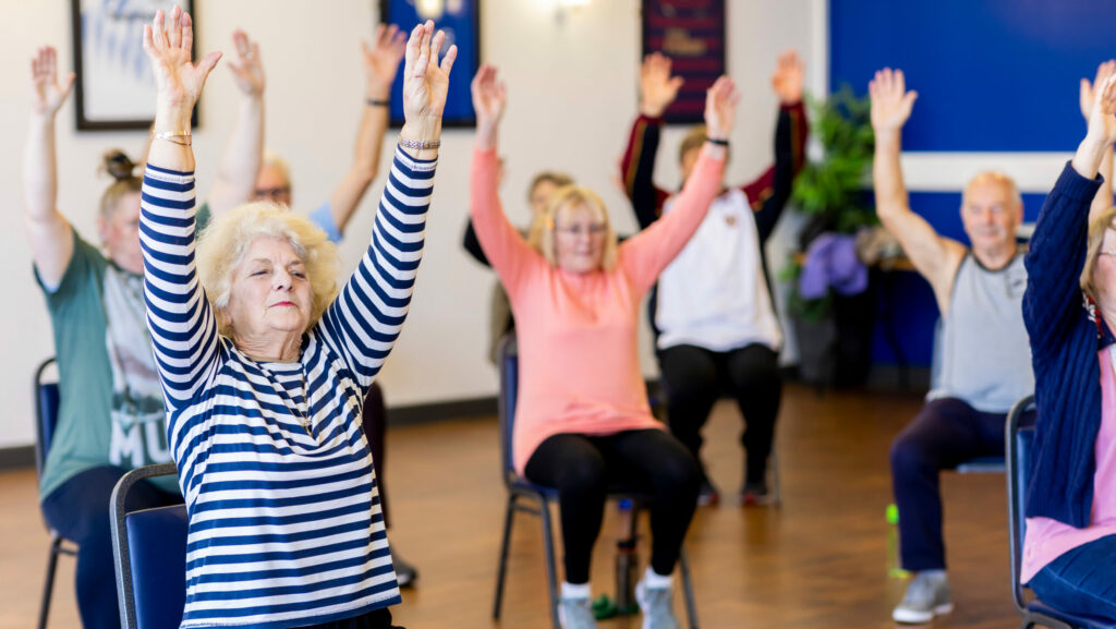 Senior:innen sitzen in einem Raum auf Stühlen und heben die Hände.