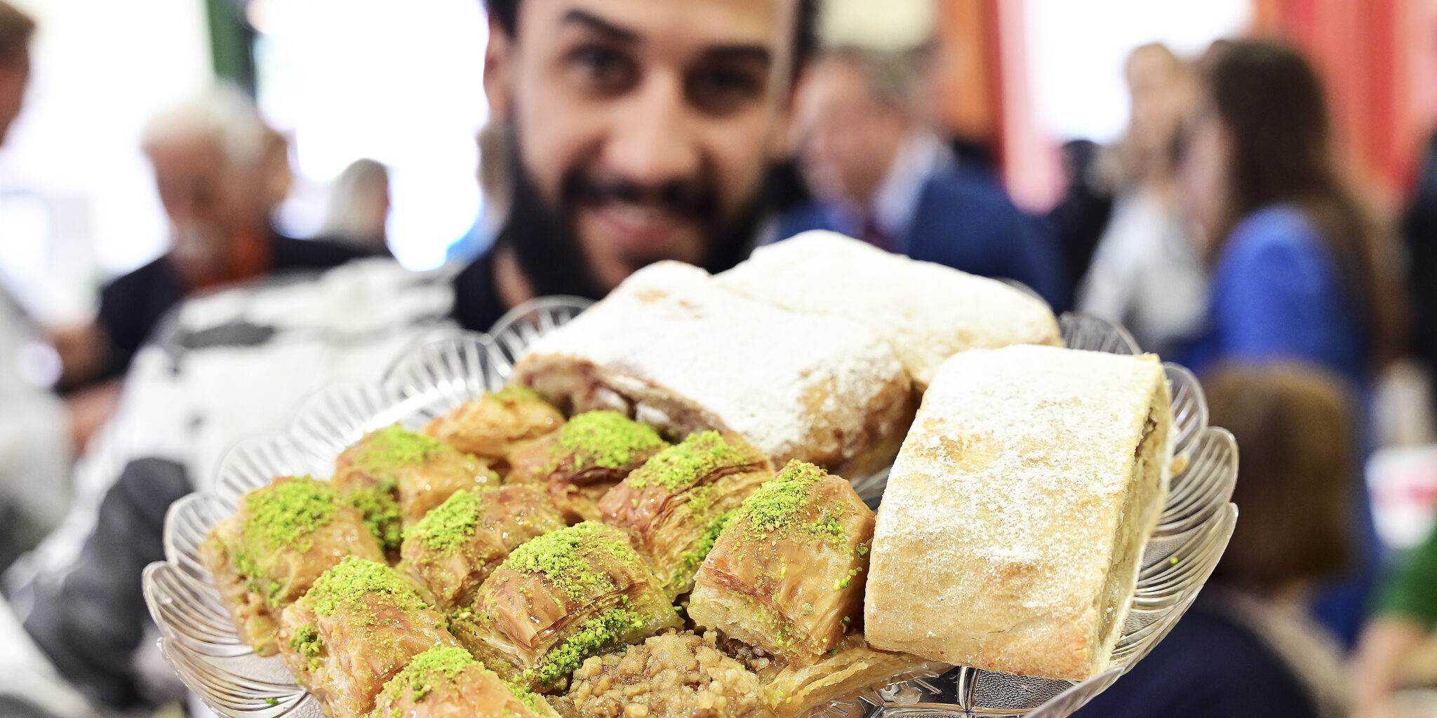 Eine Person hält ein Teller mit verschiedenen Süßspeisen in die Kamera. Vordergründig sind Stücke von Baklava zu sehen, die mit gehackten, grünen Pistazien bestreut sind. Dahinter liegen einige Stücke Strudel oder ein ähnliches Gebäck, bestäubt mit Puderzucker