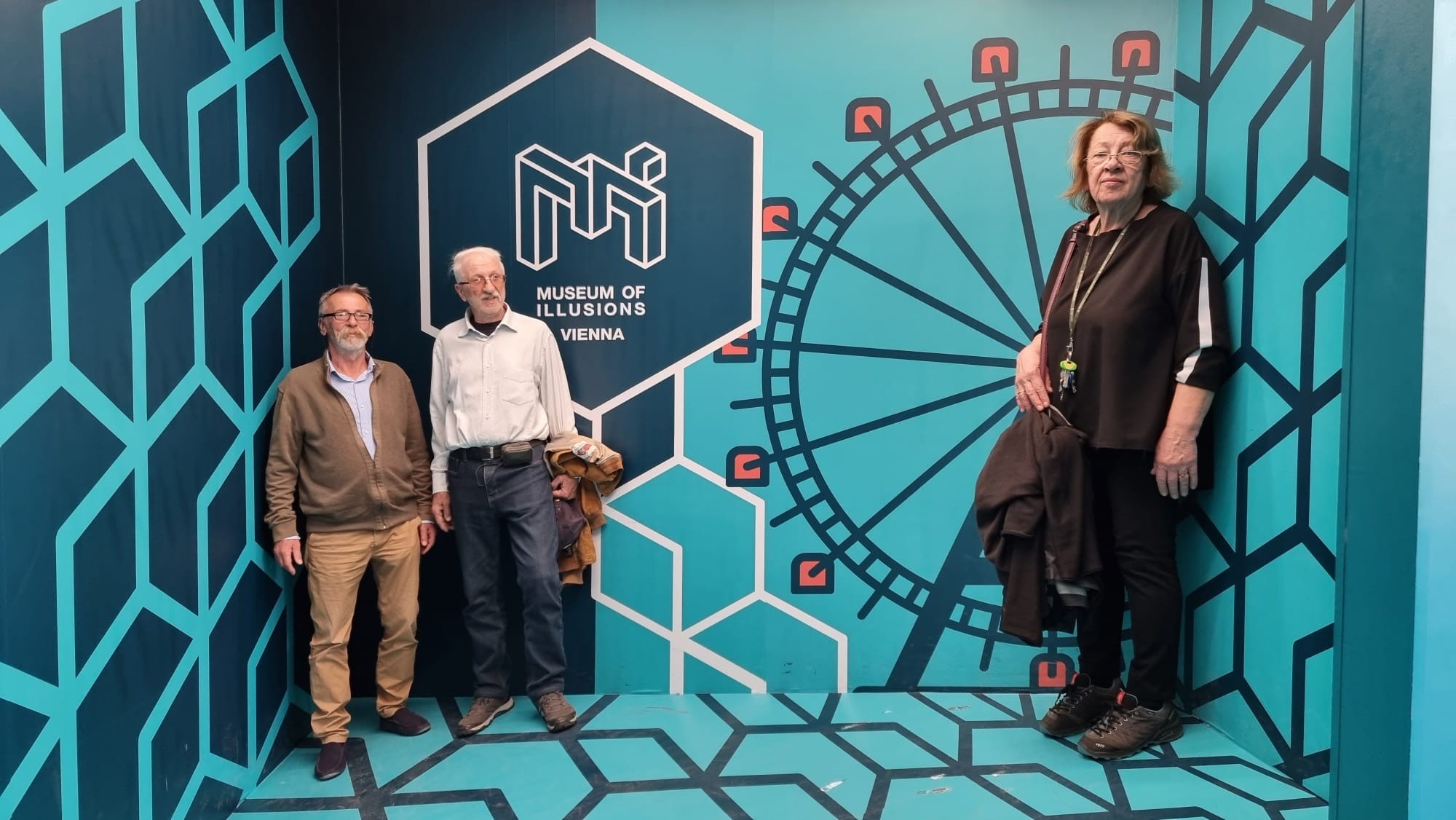 Das Bild zeigt drei ältere Personen im Museum of Illusions in Wien, wobei eine optische Täuschung den Eindruck erweckt, dass die Frau rechts deutlich größer ist als die beiden Männer links.